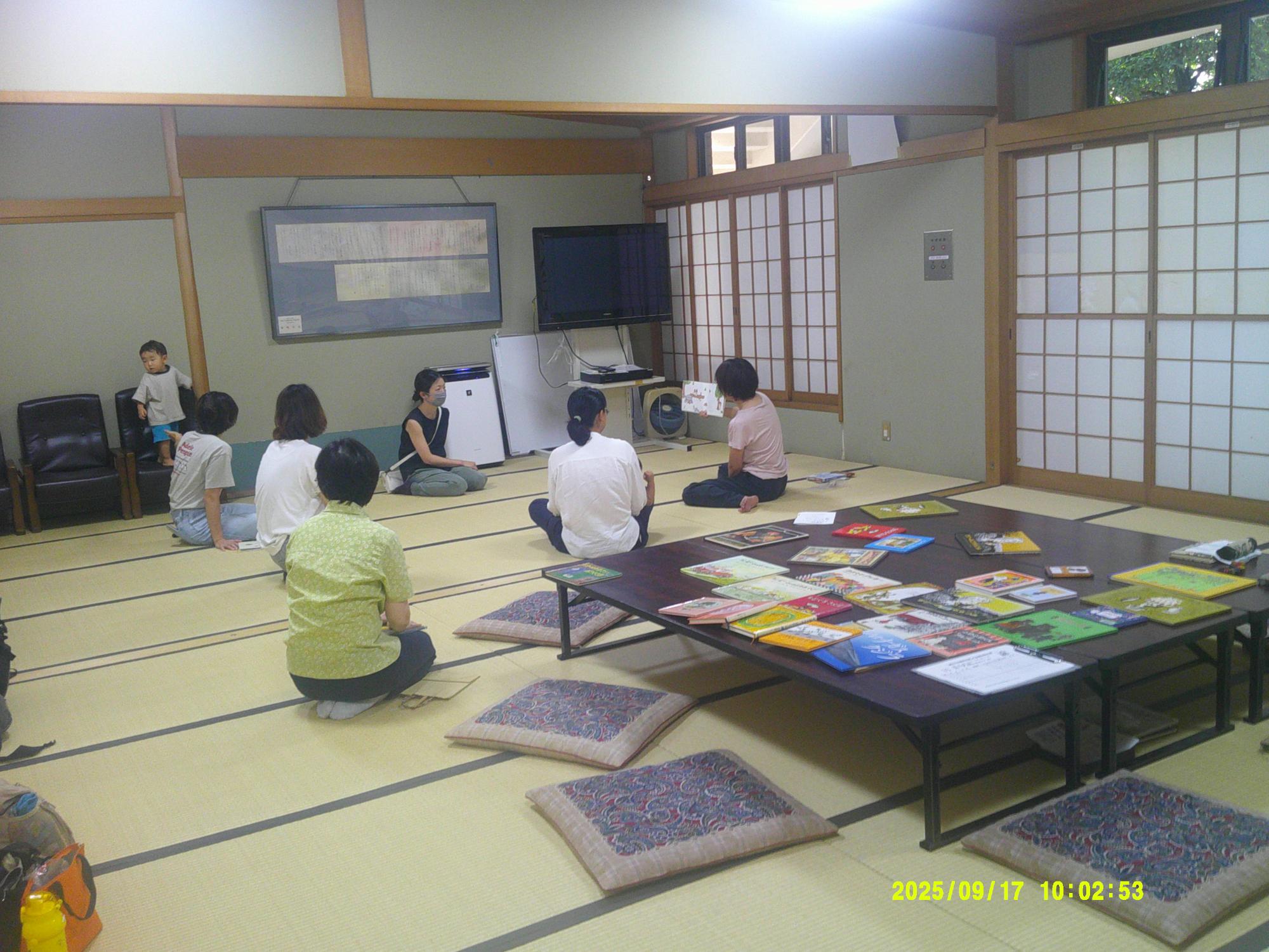 広い和室に、様々な種類の絵本が並べられており、その奥に数名の大人たちが畳の上に座って絵本の読み聞かせを行っている様子の写真