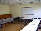 白い長机がコの字型に配置され、奥の壁には横長のホワイトボードが設置されている集会室の写コ