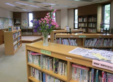 木製の書棚に多くの本が並び、手前の棚の上には紫とピンクの生け花が飾られ、新聞や雑誌が置かれている図書室の写真