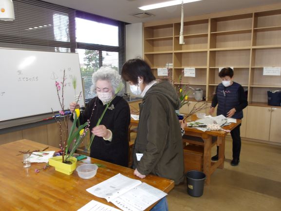 大きな机の上で講師が黄色の器に生け花を指導をしている