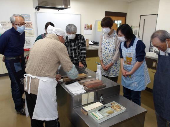 調理室で作業台を囲み講師が包丁を研いでいるところを数名で見ている