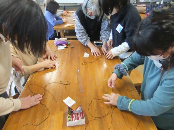 木の机の上で黒い紐を女性5人で編んでいる写真