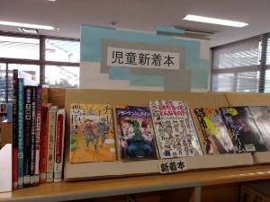 コーナーに、青と白で「児童新着本」と書かれたボードが飾られ、様々なテーマの児童書が平積みと面陳で展示されている様子を捉えた写真