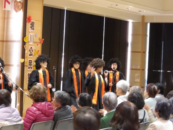 多目的ホールでかつらをかぶりオレンジの襟のついてお揃いの服を着て最後を盛り上げる人たち