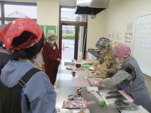 調理室で講師とアシスタントが巻き寿司を作っているところを参加者がまわりを囲んでみている