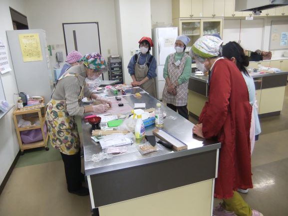 調理室で講師とアシスタントが巻き寿司を作っているところを参加者がまわりを囲んでみている