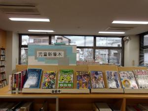 コーナーに、青と白で「児童新着本」と書かれたボードが飾られ、様々なテーマの児童書が平積みと面陳で展示されている様子を捉えた写真