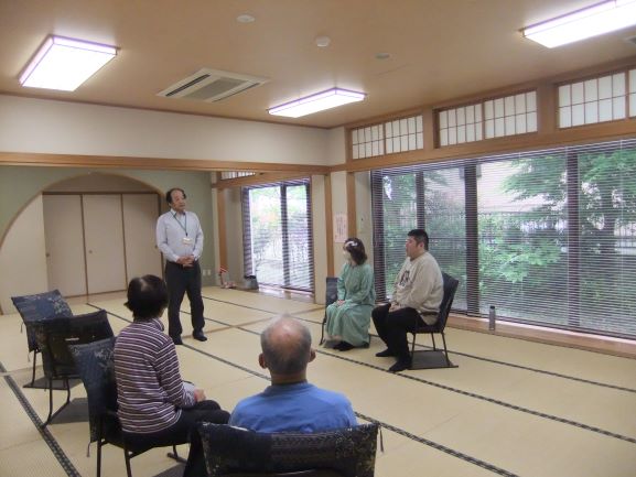 和室で男性が立って話しているのを椅子に座った大人の女性2と男性2人がいる