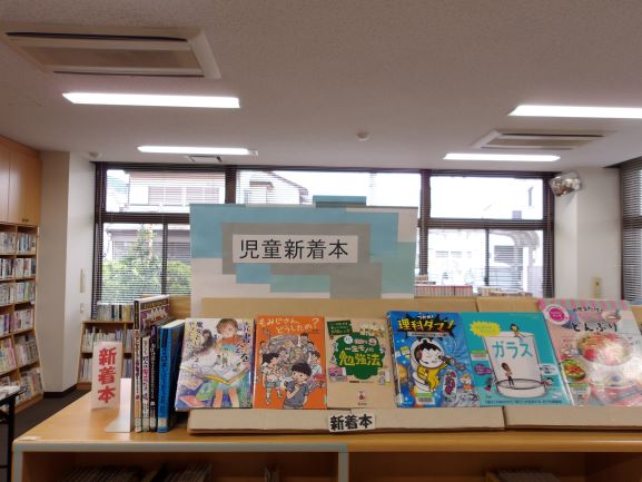 コーナーに、青と白で「児童新着本」と書かれたボードが飾られ、様々なテーマの児童書が面陳で展示されている様子を捉えた写真