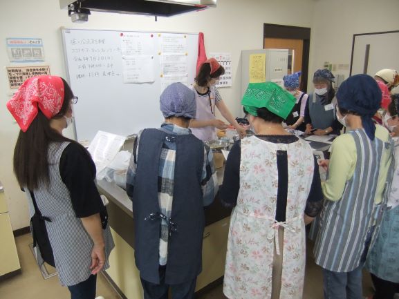 調理室で講師を囲み大人の女性が数名説明を聞いている