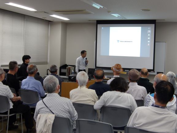 集会室で20人ほどの大人がスクリーンに映った解説を聞いている