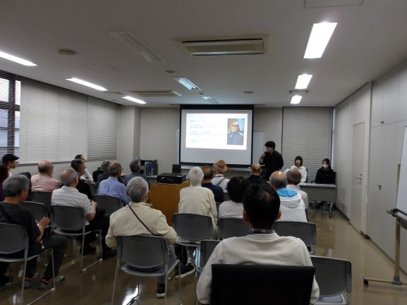 集会室で20人ほどの大人がスクリーンに映った解説を聞いている
