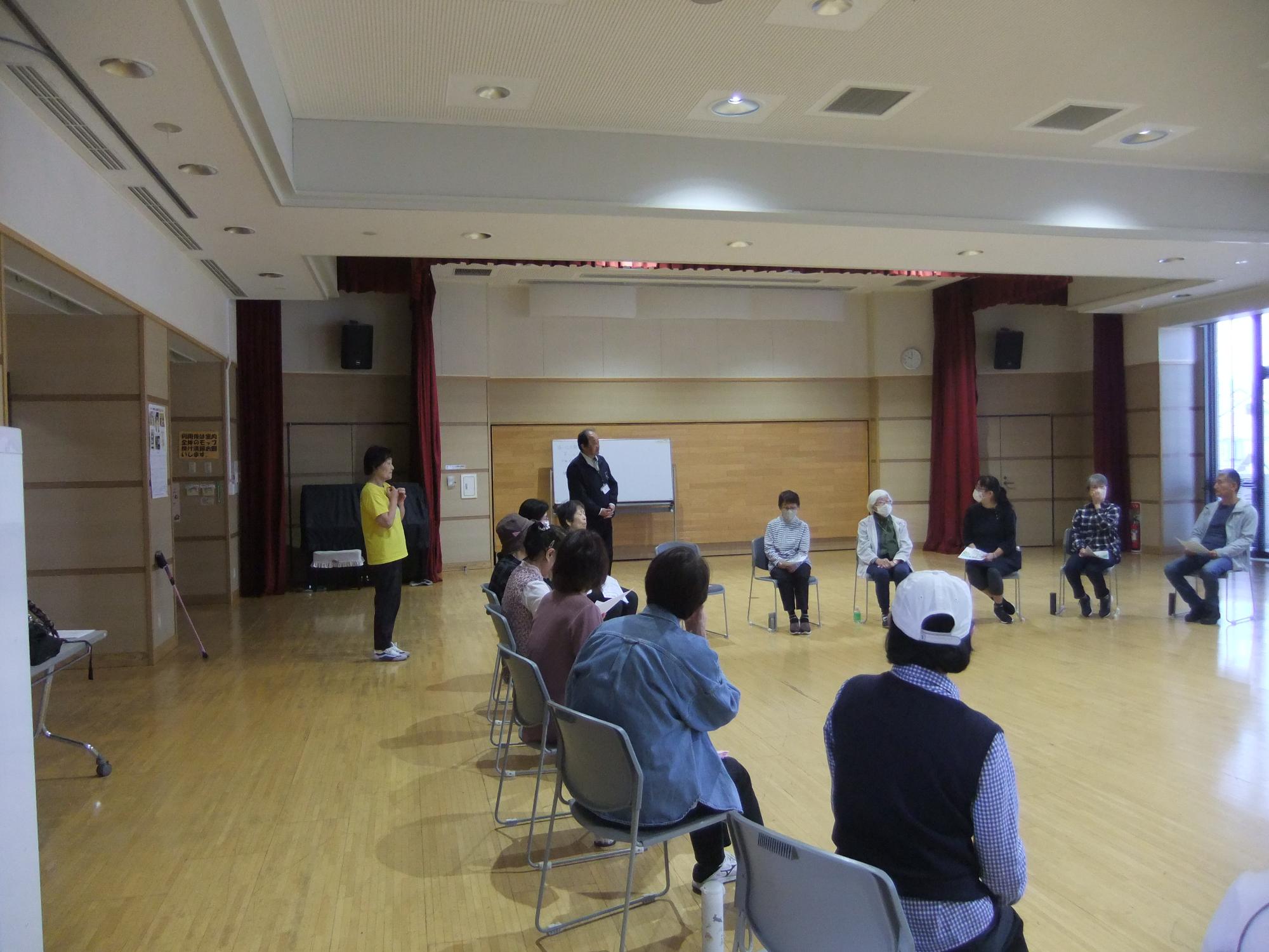ホールで多数の人が椅子に座り公民館の館長の挨拶を着ている