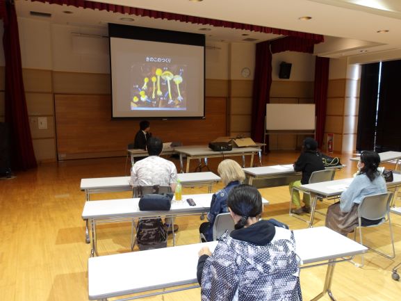 ホールで大きなスクリーンに映し出されている画像を数名の大人が見ている