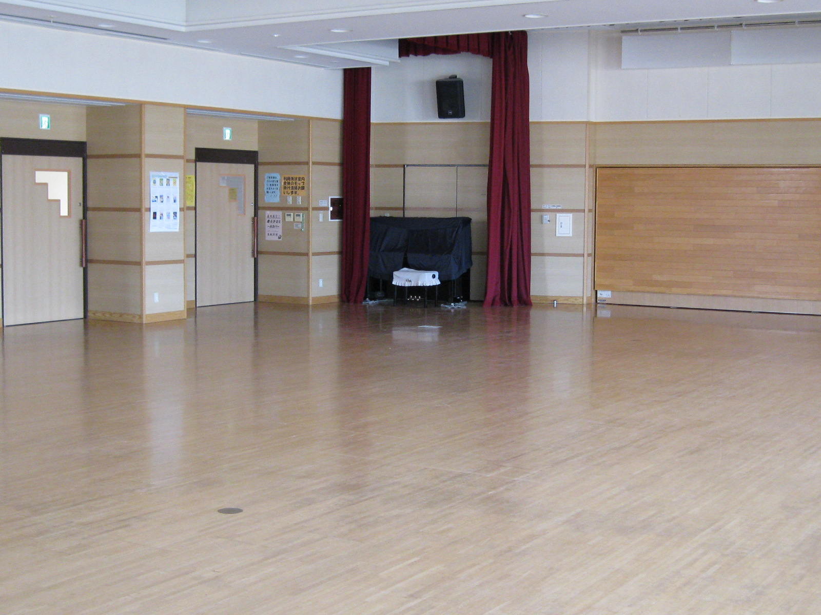 明るい木目のフローリングが広がる多目的室の内部で、前方中央には赤いドレープの幕とグランドピアノが、両脇には木目調の壁と複数の扉、掲示物が見える、イベントや集会に使われる多目的ホールの写真