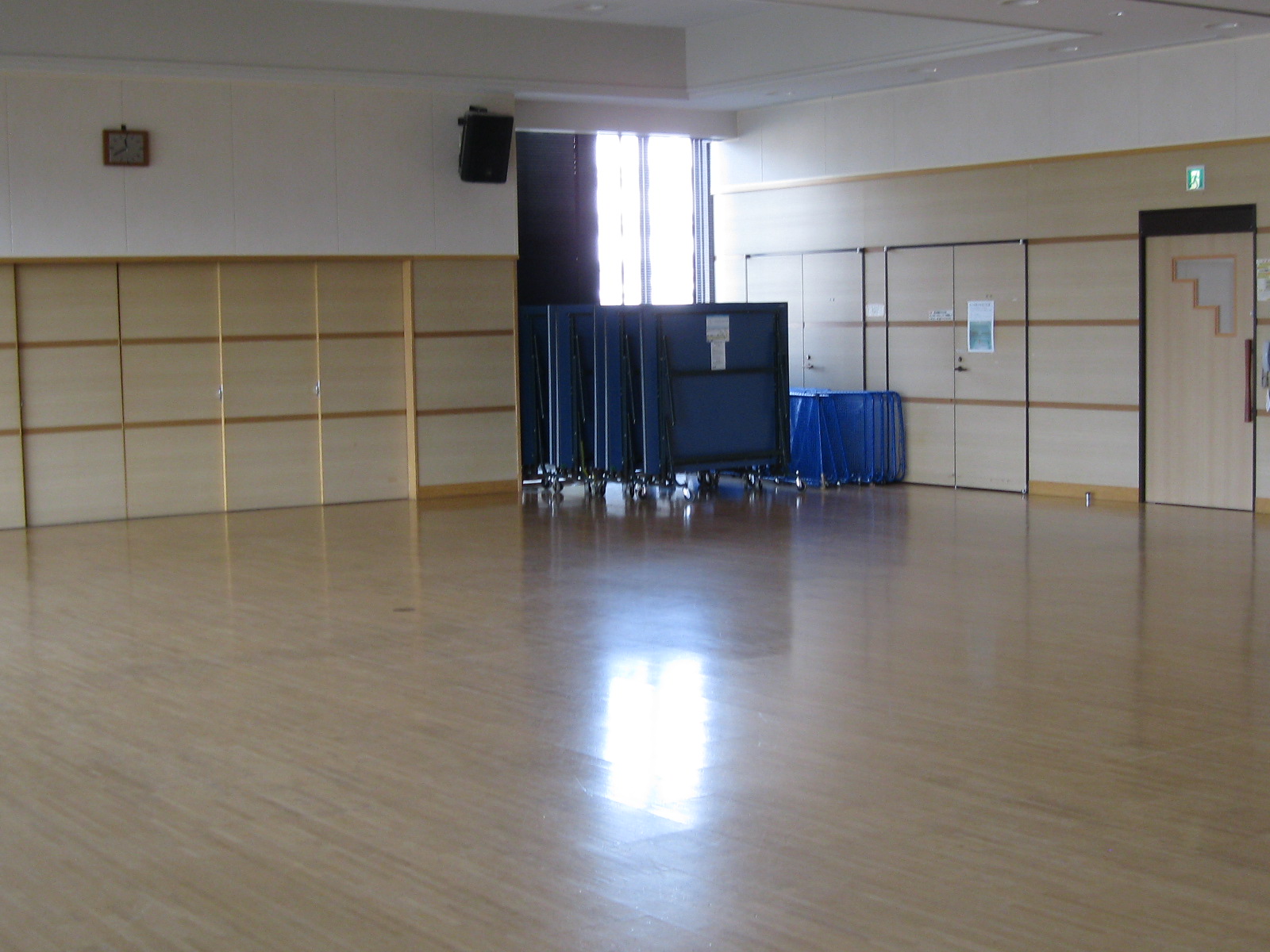 壁は白と木目調のパネルで構成され、中央の窓際には折りたたまれた複数の青い卓球台や備品がまとめて置かれている多目的ホールの写真