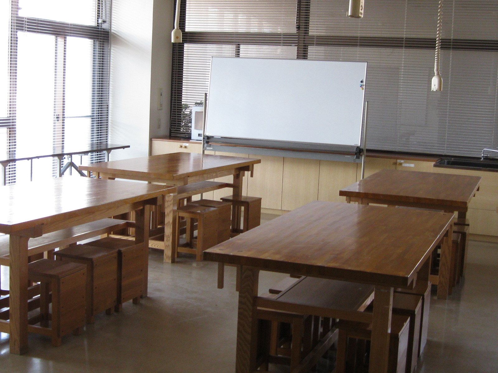 ブラインド付きの大きな窓から光が差し込む明るい部屋の内部で、重厚な木製の長机が数台と、その下に収納できる角形の木製スツールが配置され、正面奥には移動式のホワイトボードと収納棚が見え創作活動室の写真