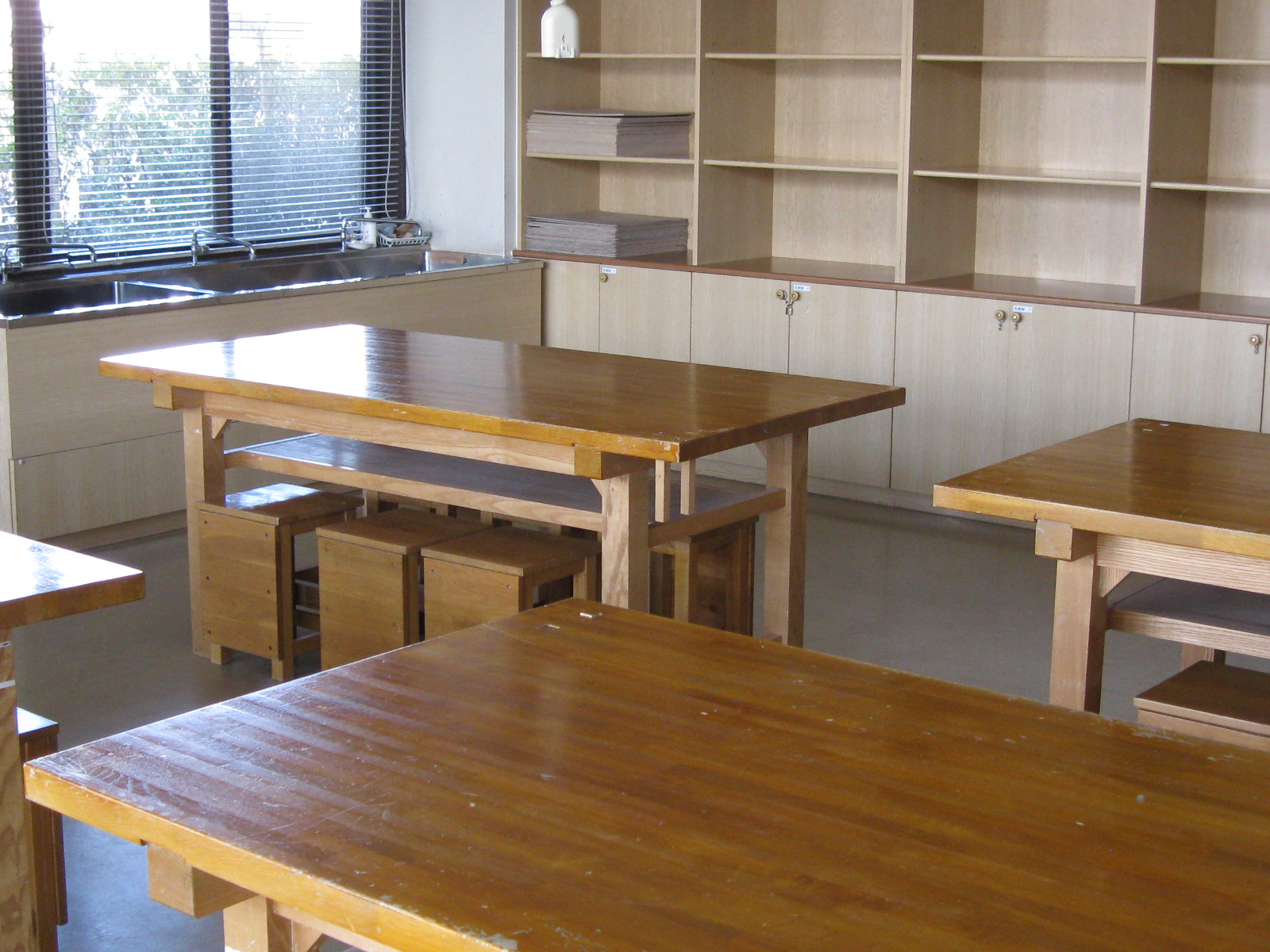 光沢のある木製の大型作業台が複数配置され、奥の壁際にはステンレス製のシンクと、収納棚とキャビネットが備え付けられている創作活動室の写真