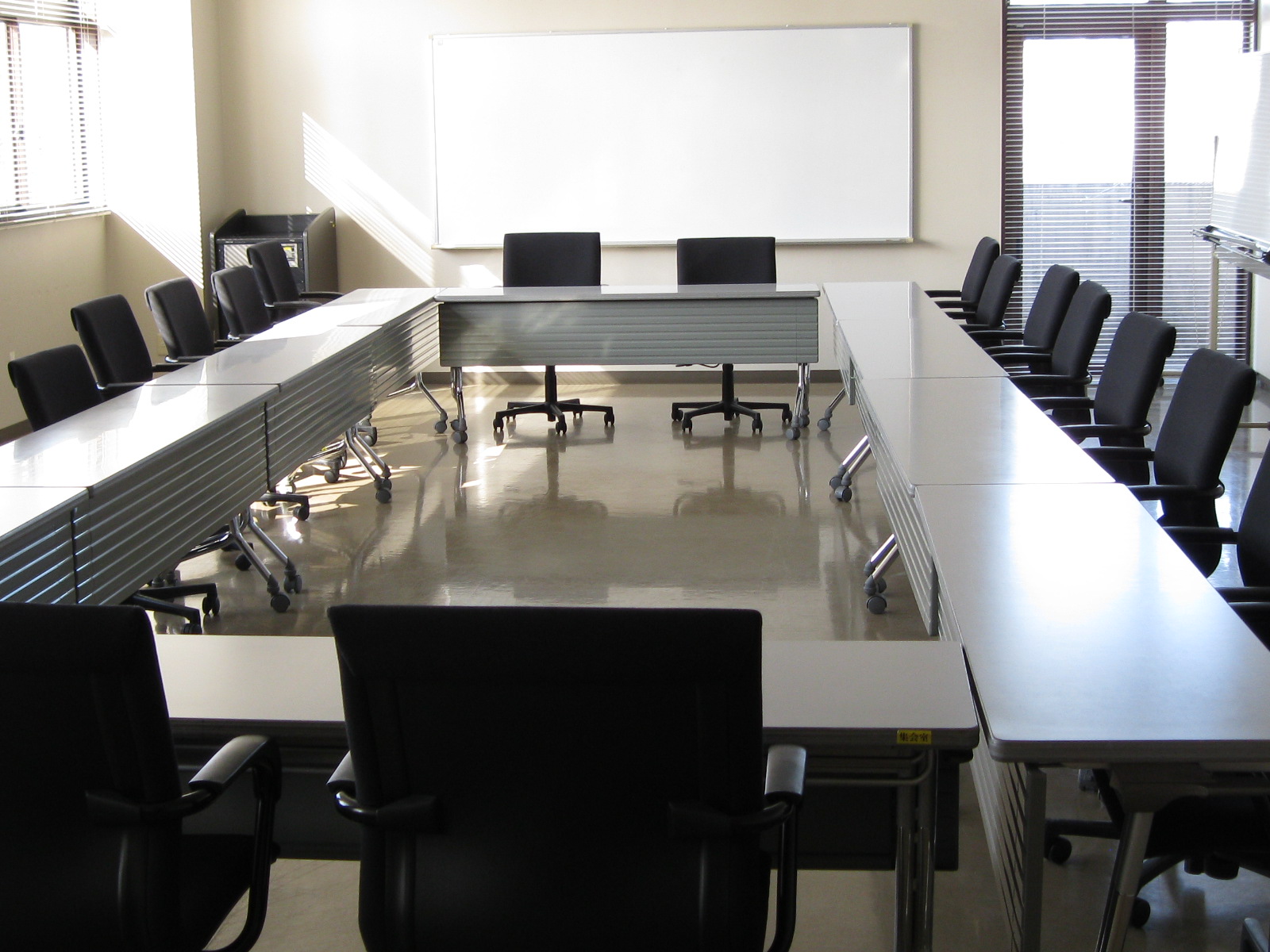 長机と黒い事務椅子がコの字型に配置され、部屋の正面奥の壁には大きなホワイトボードが設置されている集会室の写真