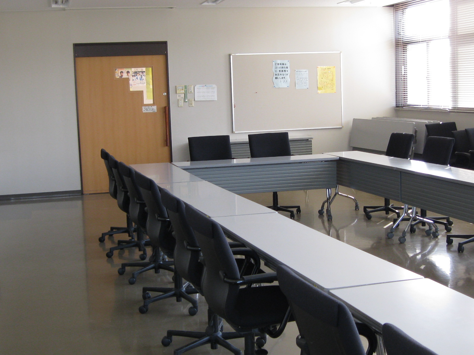 長机と黒いキャスター付きの椅子が右側に長く伸びるコの字型に配置され、奥の壁には掲示板、左奥には木目調の扉と窓がある集会室の写真