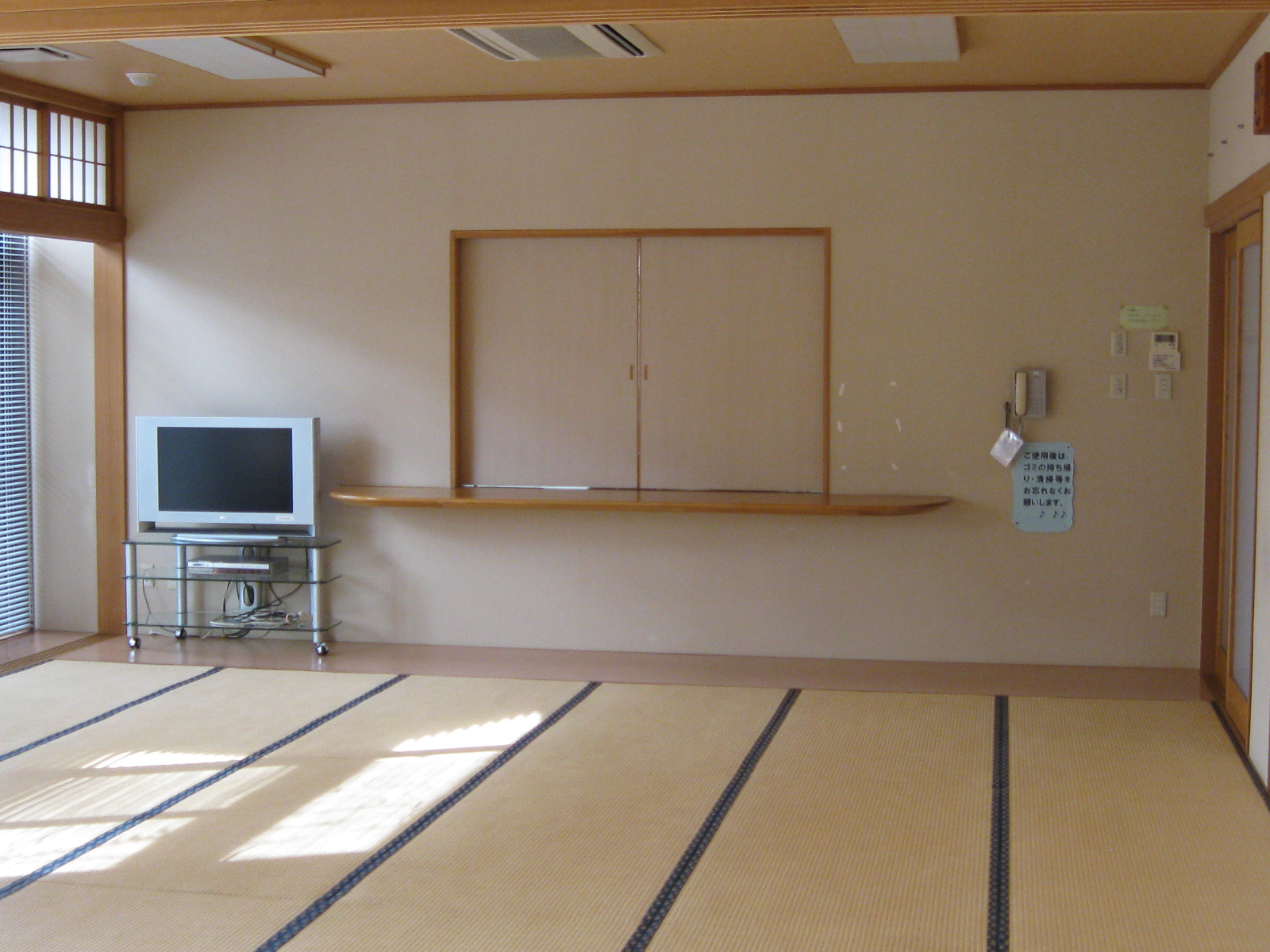 畳敷きの床とクリーム色の壁を持つ和風の休憩室で、奥の壁には障子のような窓と、その下に木製のカウンターがあり、左側にはテレビが置かれたガラスのテレビ台がある、和室の写真