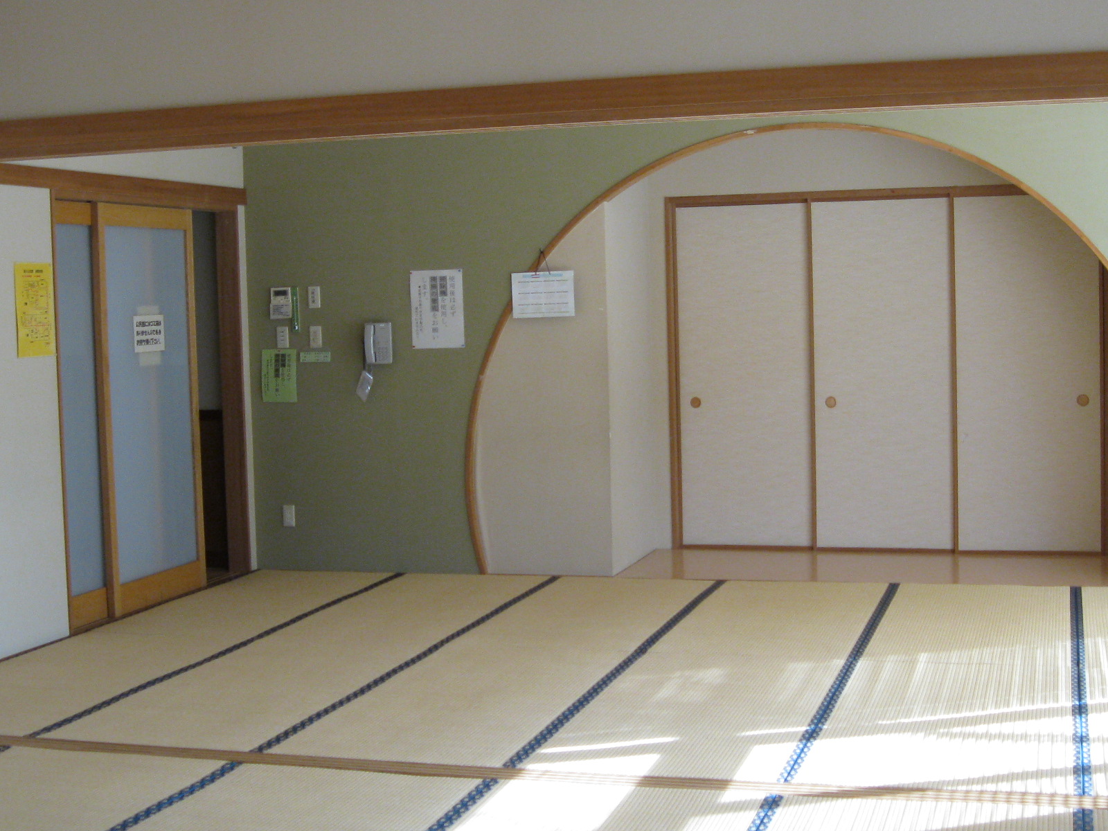 畳が敷かれた和室の内部で、右奥に障子戸の押し入れがあり、その手前はカーブを描いた壁に囲まれ、左側には掲示物が貼られた薄緑の壁と、ガラス入りの引き戸がある和室の写真