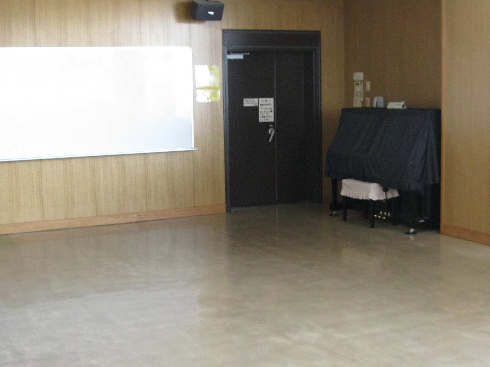 木目調の壁が特徴的な部屋の内部で、壁面には大きなホワイトボードが設置され、中央には黒い扉、右側には黒いカバーがかけられたアップライトピアノが置かれている音楽室の写真