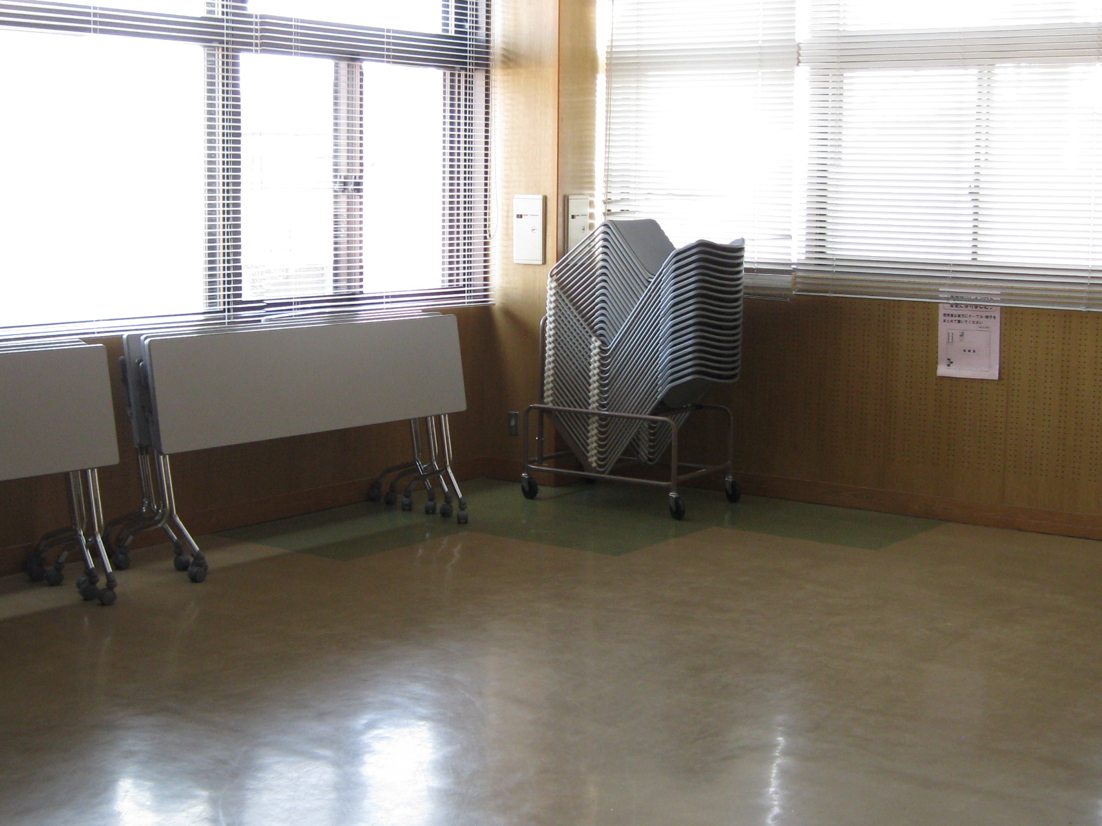ブラインド越しに光が差し込む大きな窓の前で、キャスター付きの台に積まれた大量の折りたたみ椅子と、折りたたまれて立てかけられた会議用の長机が整然と保管されている音楽室の写真