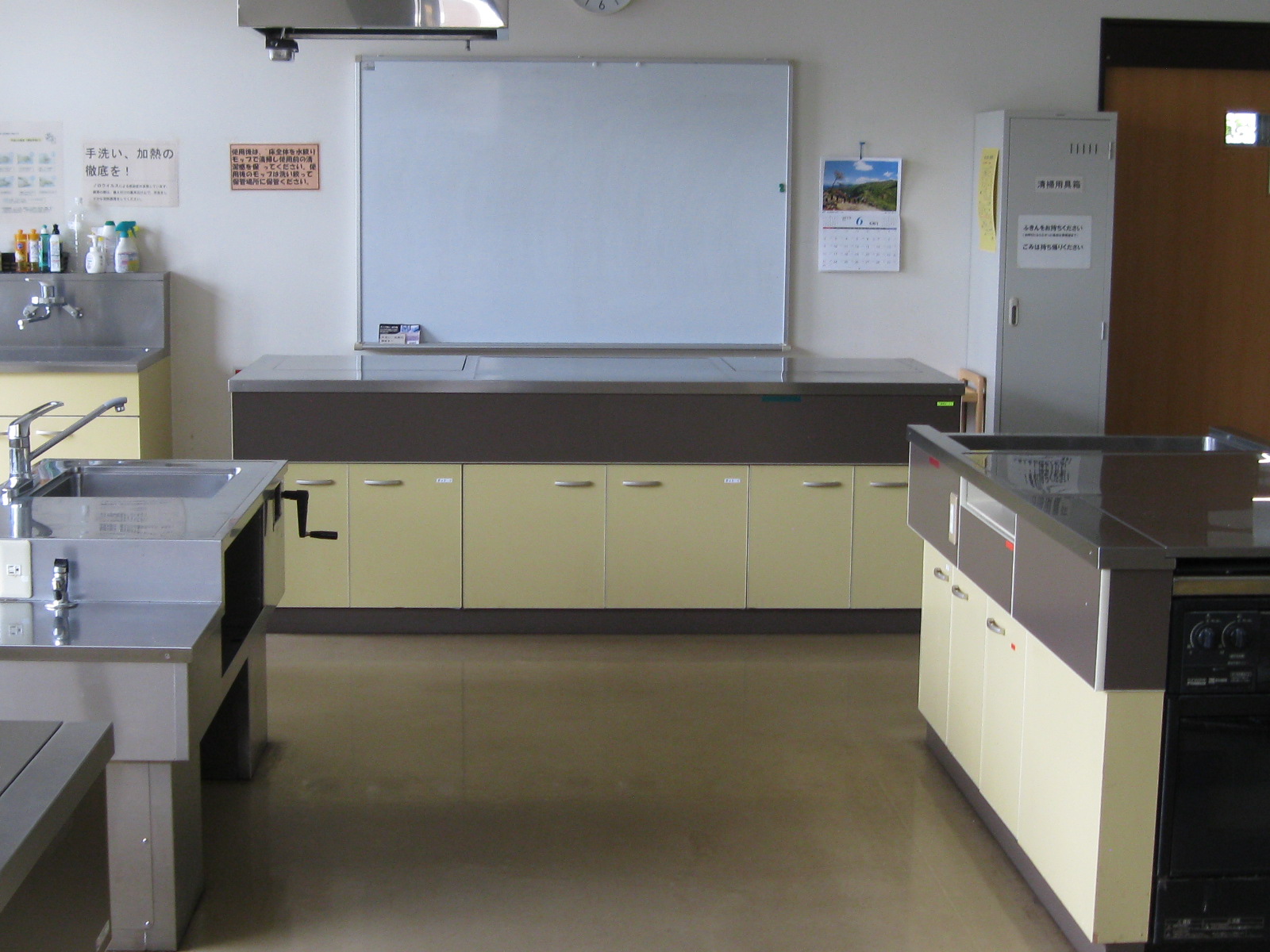 シンク付きのステンレス製調理台が複数置かれた清潔感のある調理室の風景で、中央には黄土色のキャビネットを備えたカウンターと、その上に大きなホワイトボード、壁には注意書きとカレンダーが貼られている調理室の写真