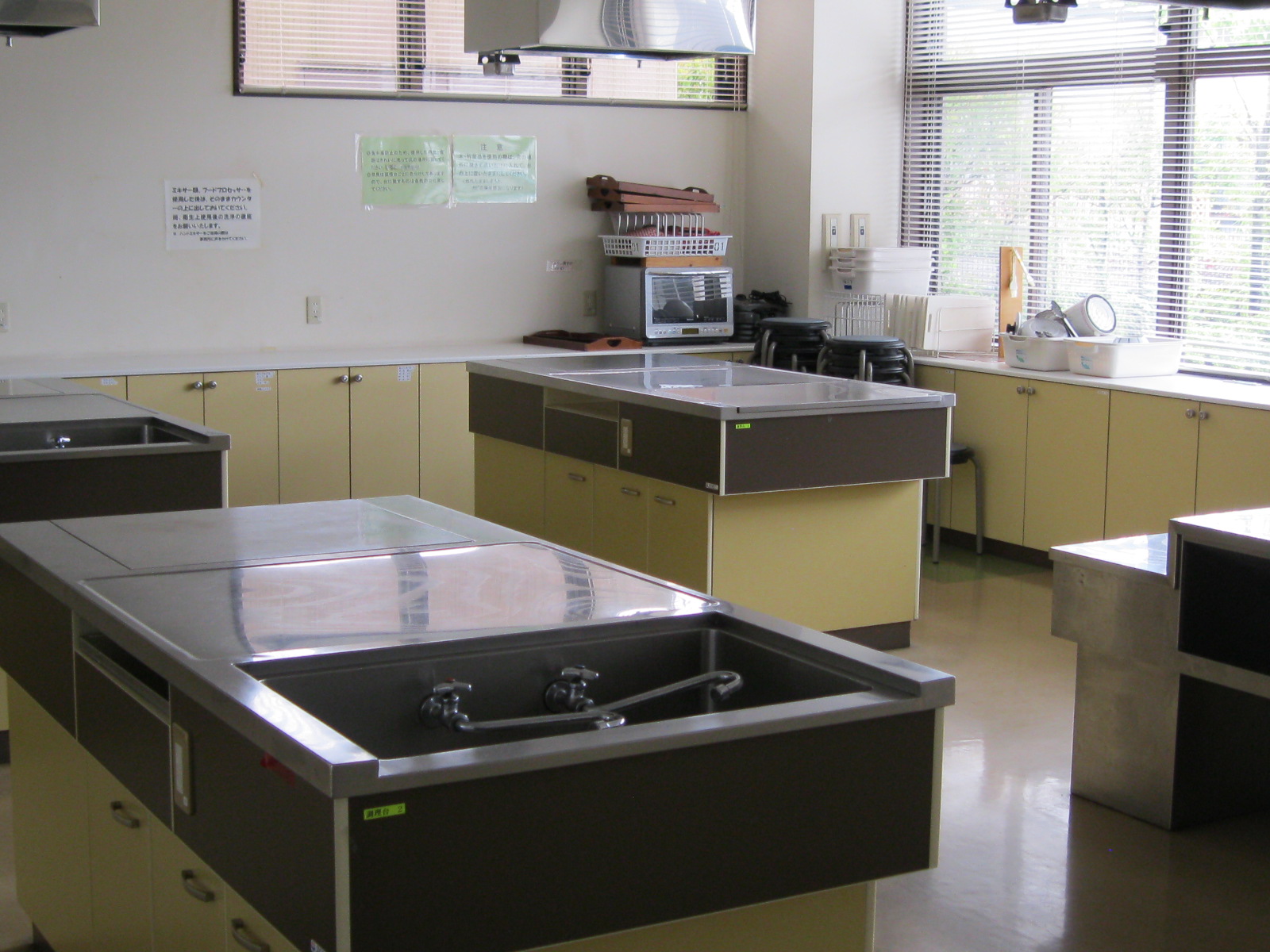 ブラインド付きの大きな窓から光が入る調理実習室の内部で、シンクと作業スペースを備えた茶色と黄土色のキャビネットの調理台が複数設置されており、壁には注意書きが貼られ、奥には電子レンジや食器類などの備品が置かれている調理室の写真
