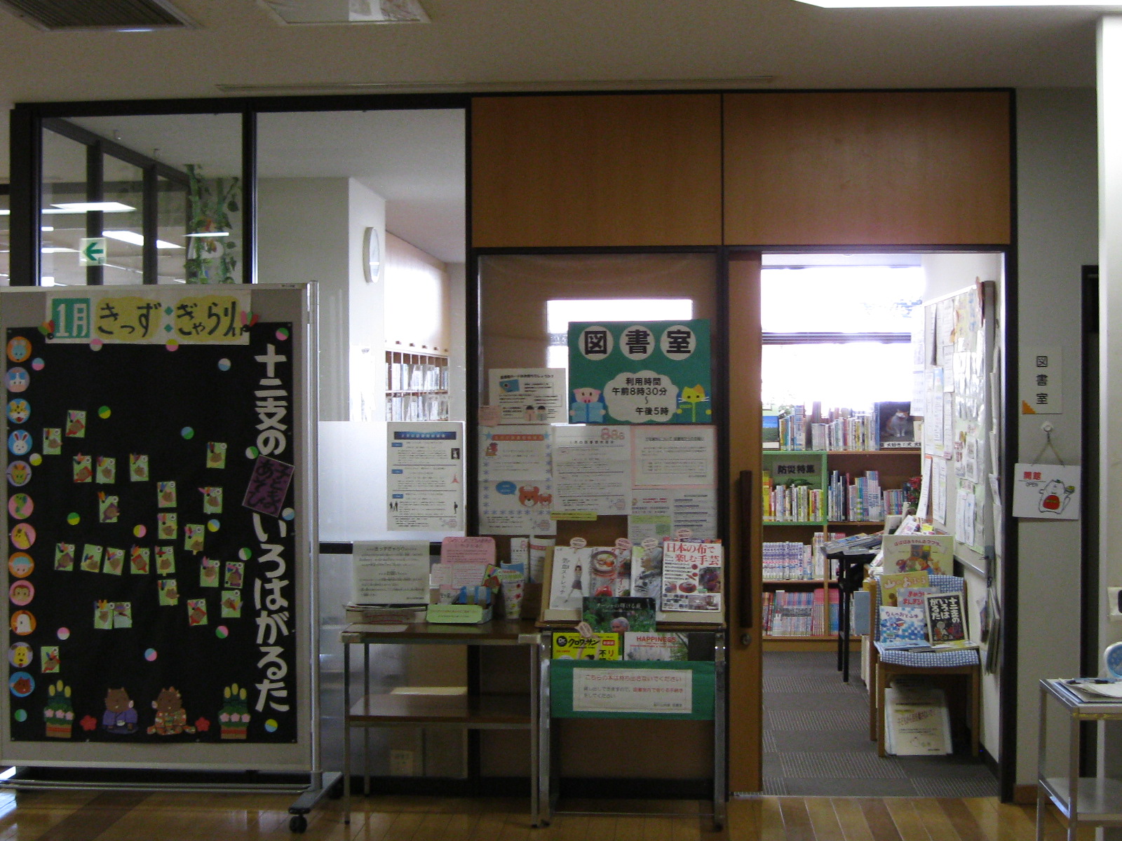 学校または地域の図書館の入口付近を写した写真で、左側には装飾された黒板の掲示板、右側には「図書室」の案内表示と共に、多数のポスターやパンフレット、書籍が所狭しと並べられた受付前のスペースがある図書室入り口付近の写真