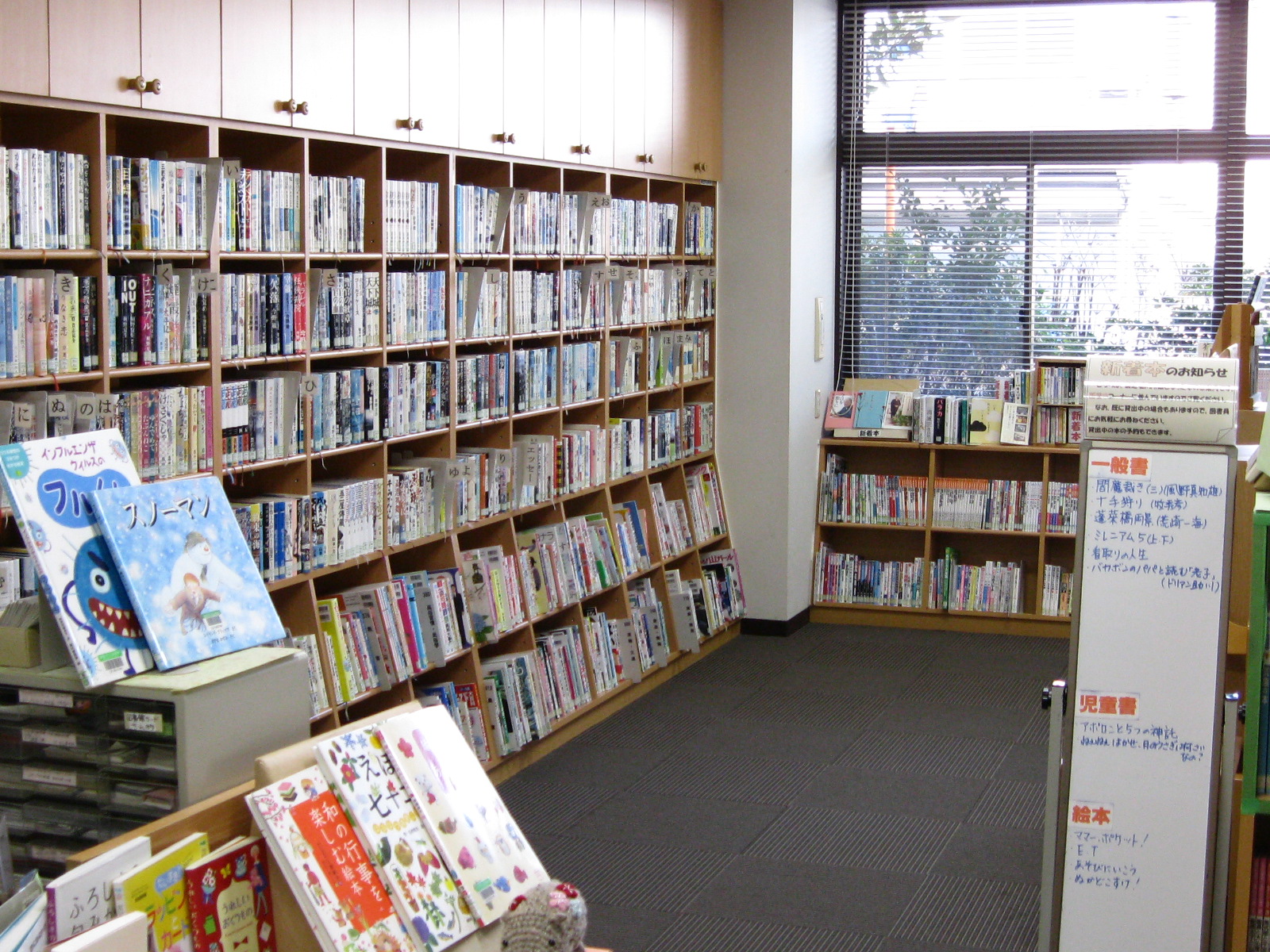 木目調の壁一面に本棚が設置され、小説や実用書などが大量に並んでおり、手前には児童書や絵本が展示され、右側にはブラインド付きの大きな窓がある、明るい図書室の写真