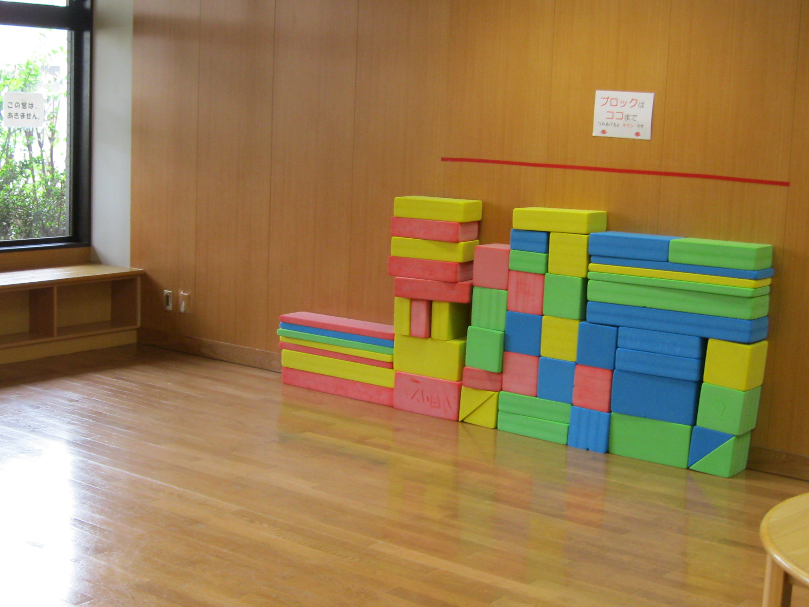 木目調の壁と床が特徴的な子供向けの遊び場の一角で、色とりどりの柔らかい大型積み木が壁際に積み重ねられ、左奥には窓と低い棚、右奥の壁には「ブロックはココまで」という表示がある児童室の写真