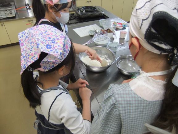 白衣とバンダナを身につけた子供たちが、流し台のある調理室で、エプロン姿の女性講師からお菓子か料理の作り方を教わっている様子の写真