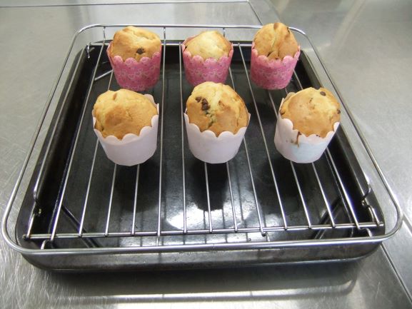調理場のステンレスの作業台の上に置かれた天板の網の上に、焼きあがったばかりのチョコレートチップ入りのカップケーキが6つ並んでいる様子の写真