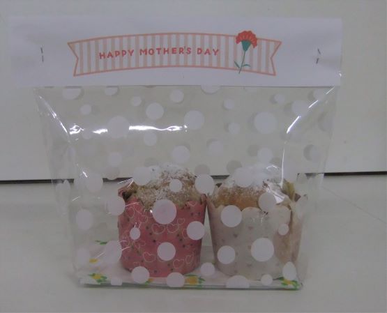 ピンク色と水玉模様のカップケーキ型に入ったマフィンが2つ、ドット柄の透明な袋にラッピングされており、上部には「HAPPY MOTHER'S DAY」のメッセージが書かれた写真