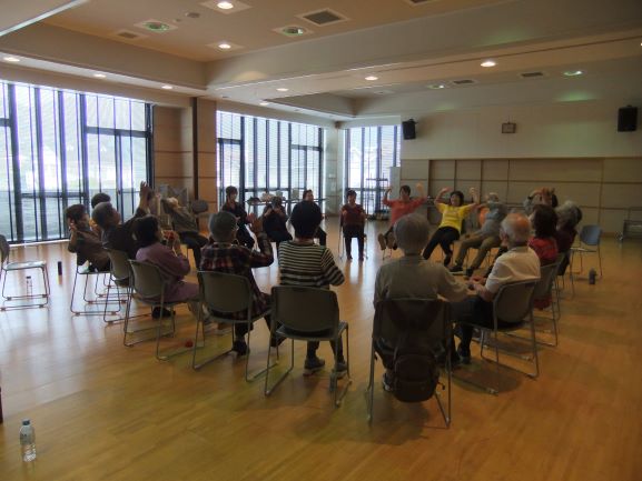 黄色いシャツを着た女性の佐久間さんが椅子に座った高齢者たちと輪になって体操をしている様子の写真