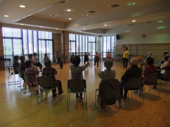 黄色いシャツを着た女性の佐久間さんが、体育館のような広々とした部屋で、椅子に座った高齢者たちと輪になって体操をしている様子の写真