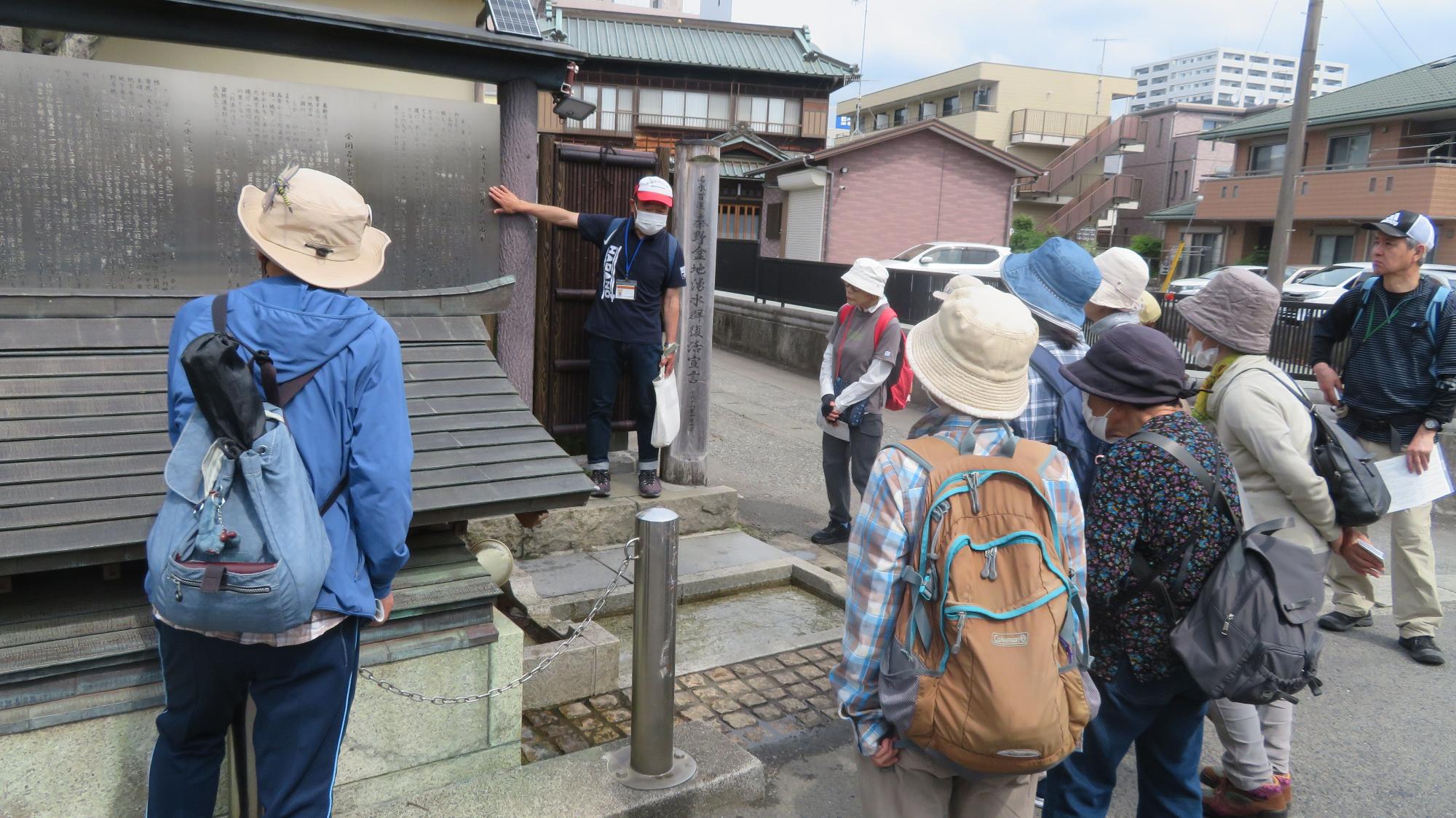 数人の高齢者グループが、青い服と白い帽子を被った男性の説明を聞きながら、町の古い建物にある石碑を囲んで見ている様子の写真