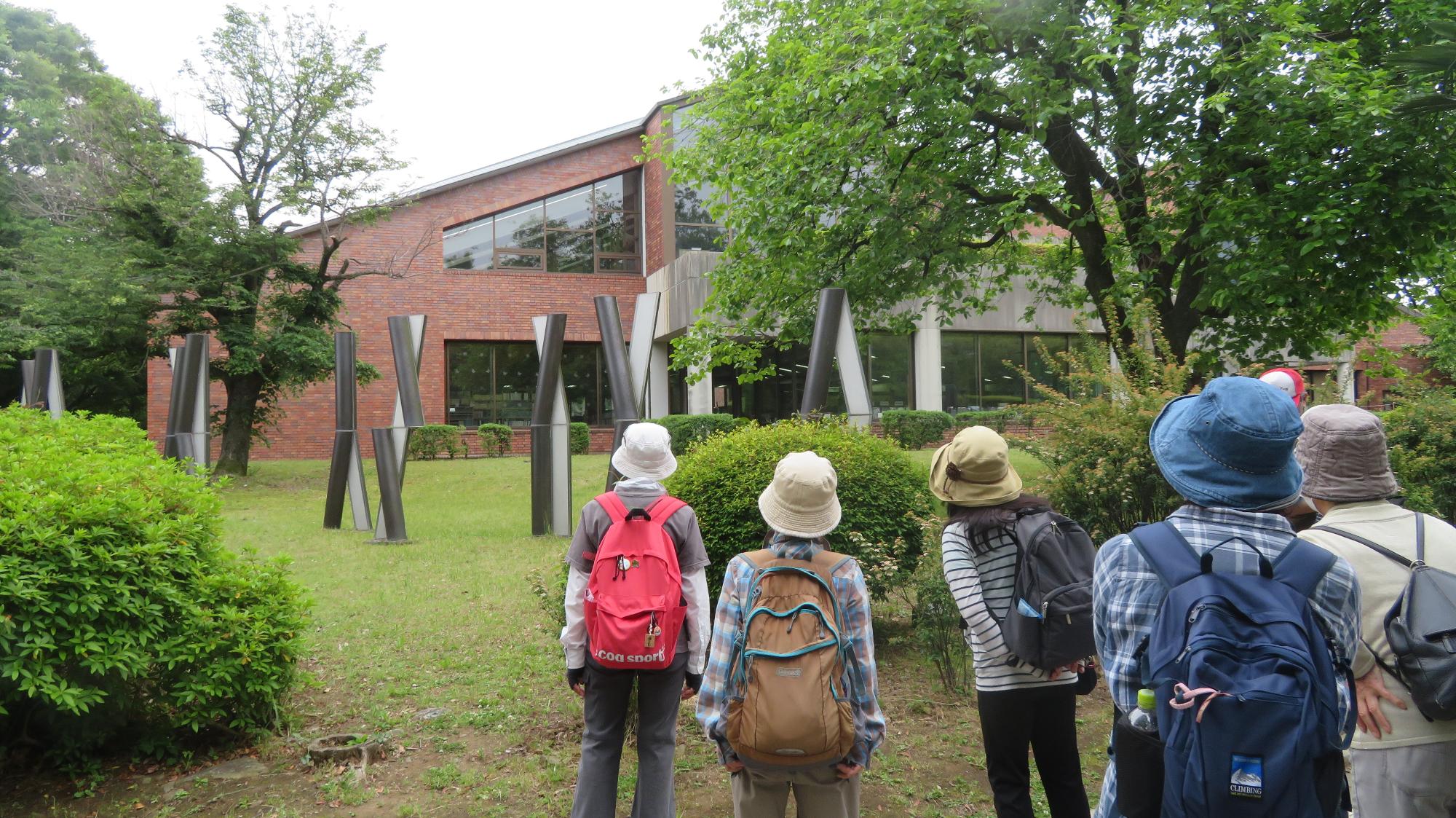赤煉瓦の建物と緑豊かな庭園を背景に、複数の人々が、リュックを背負って建物の方を眺めている様子の写真