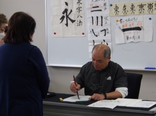 書道教室で細川さんが机に向かって筆を運び、生徒の一人がその手元を熱心にのぞき込んでいる様子の写真