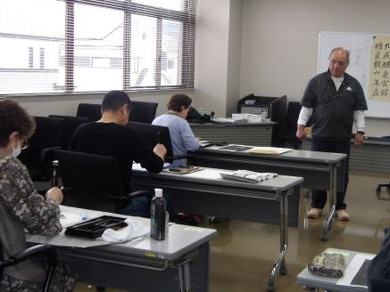 教室内で、細川さんが机の間を歩きながら生徒の様子を見ており、複数の生徒が各自の席で書道や絵画の練習をしている様子の写真