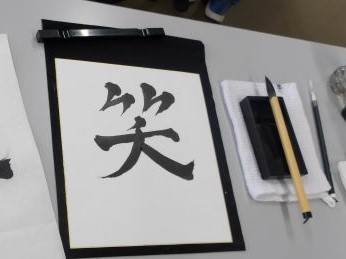 黒い表装の半紙に筆で大きく「笑」という漢字が書かれており、その横には硯や筆が置かれた書道教室の机を写した写真
