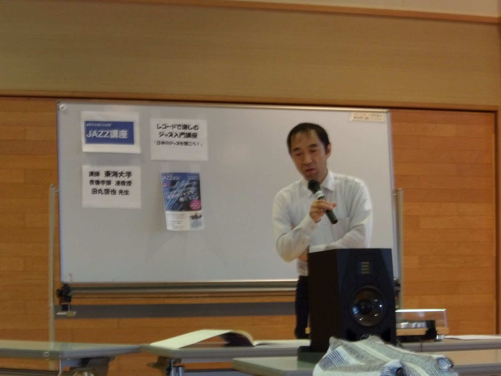 ホワイトボードの前に立ち、マイクを片手に話している田丸さんと、ボードに貼られた「JAZZ講座」と書かれた貼り紙を写した写真