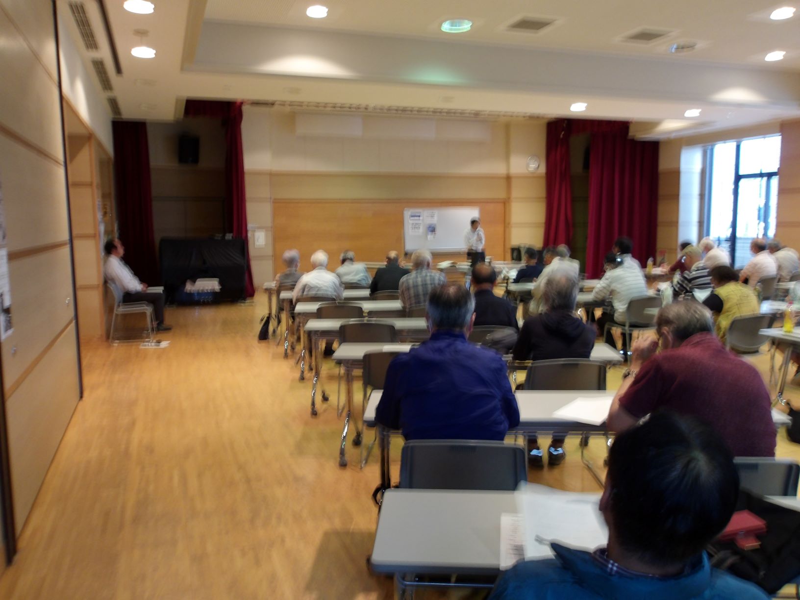 多くの聴衆が教室で机に向かって座っており、壇上の講師が説明している様子を、部屋の奥から写した写真