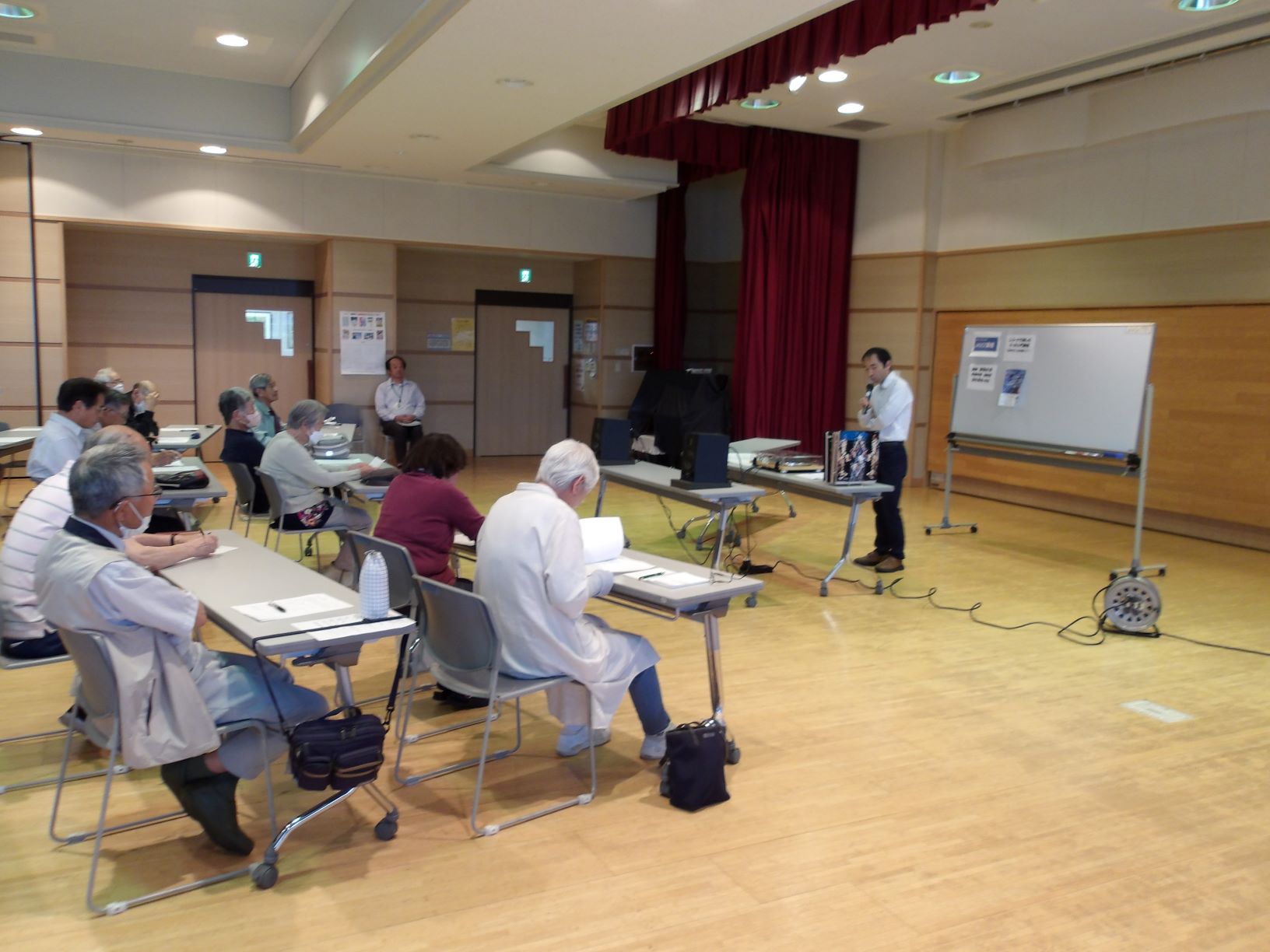 床にケーブルが這わされた広い部屋で、複数の高齢者が机に座って講師の話を聞いている、JAZZ講座の様子の写真