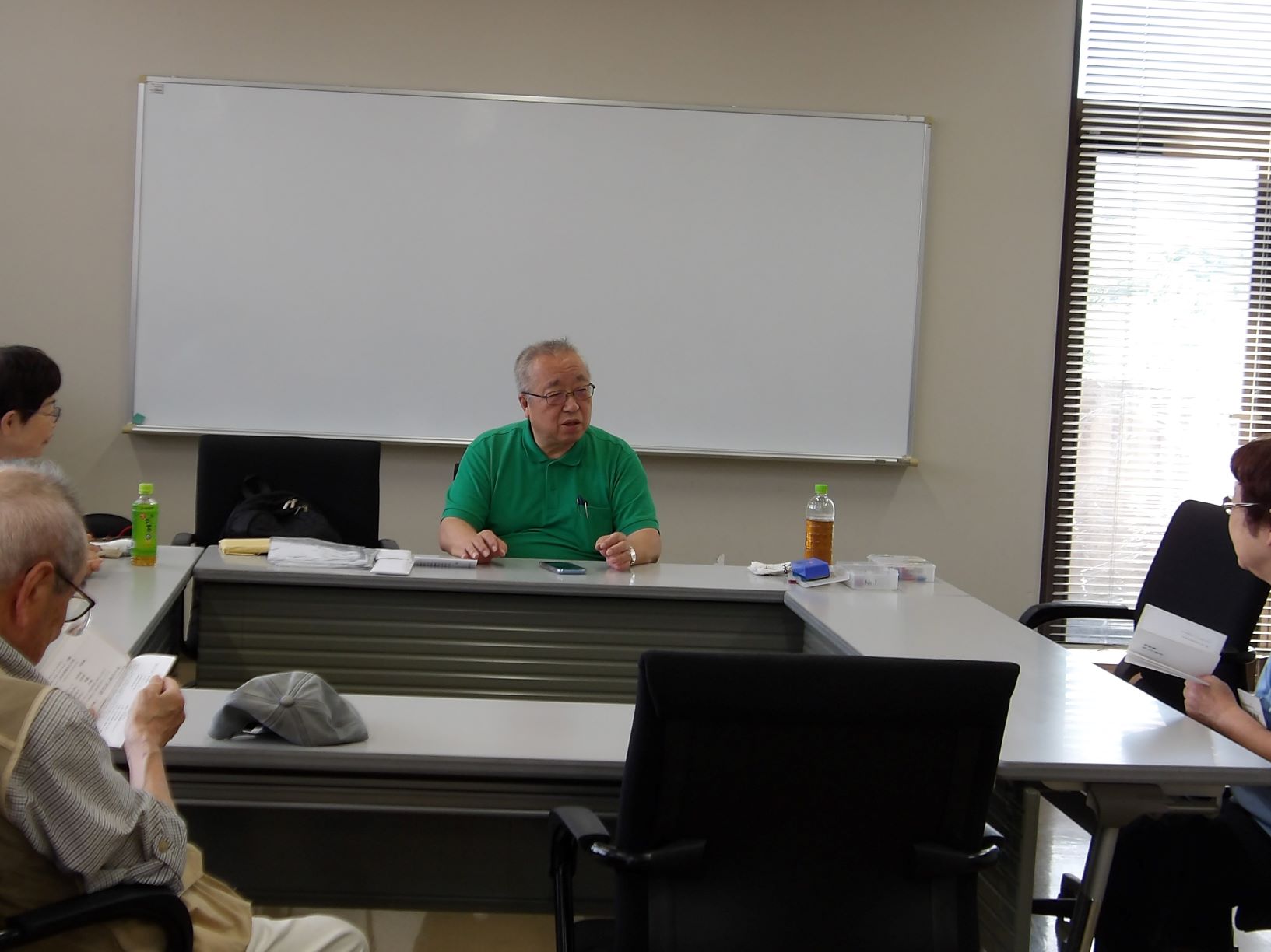 緑色のポロシャツを着た男性が会議室のテーブル席に座り、身振り手振りを交えながら他の参加者に向けて話している様子の写真