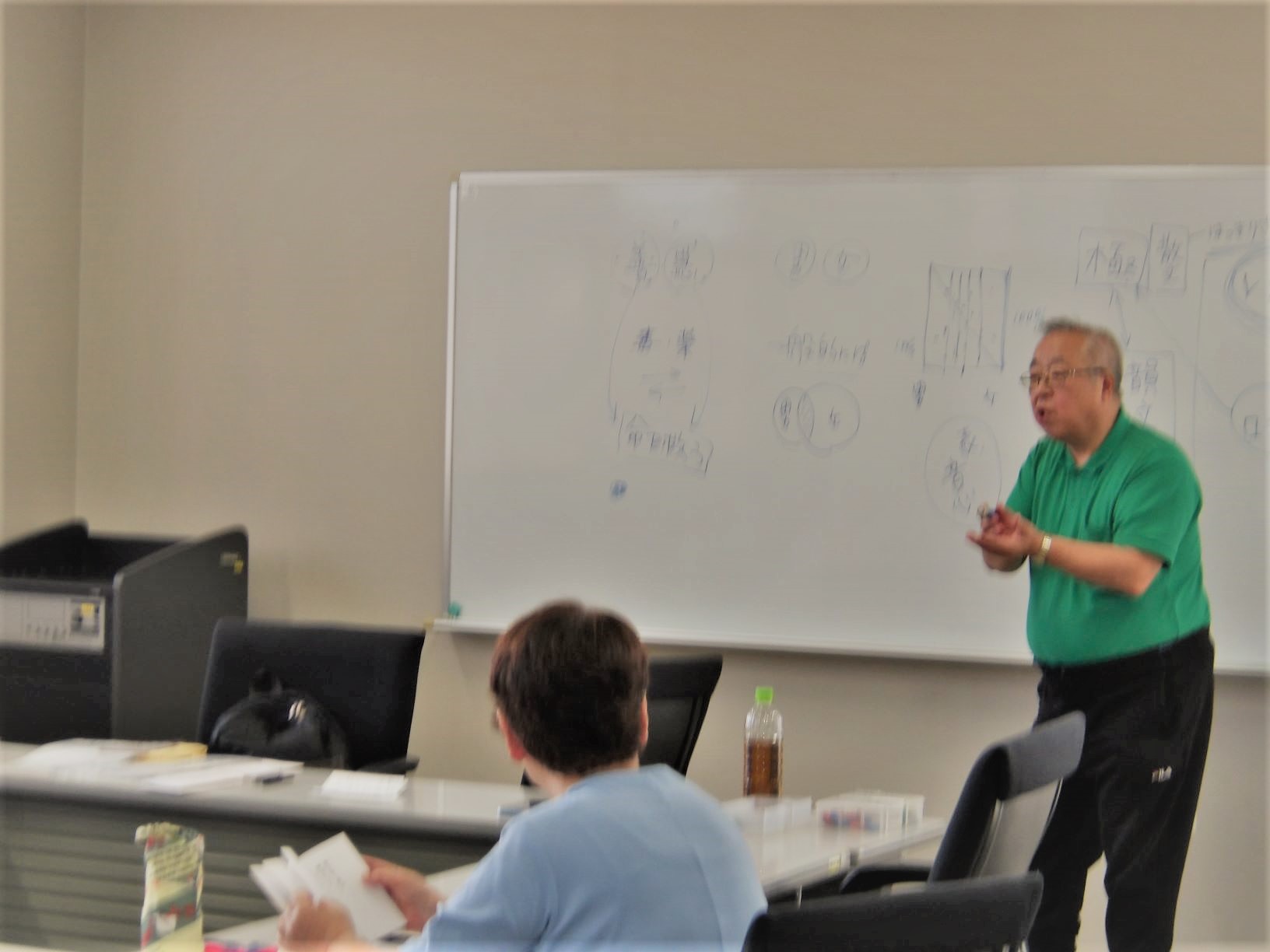 緑色のポロシャツを着た男性が、ホワイトボードを背に、身振り手振りを交えながら聴衆に向かって熱心に説明をしている様子の写真