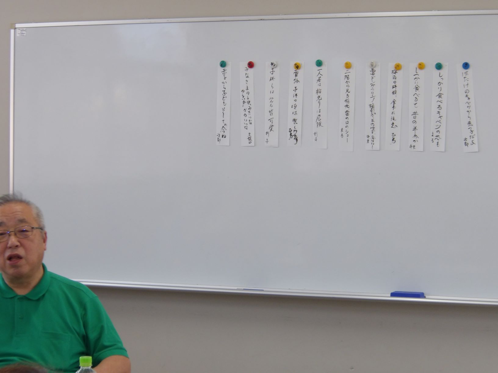 眼鏡をかけた男性が緑色のポロシャツを着て、ホワイトボードに向かって座っている様子の写真