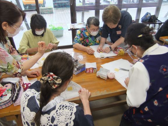 数名の女性が、マスクを着用してひとつのテーブルを囲み、裁縫道具を使って熱心に手芸に取り組んでいる様子の写真